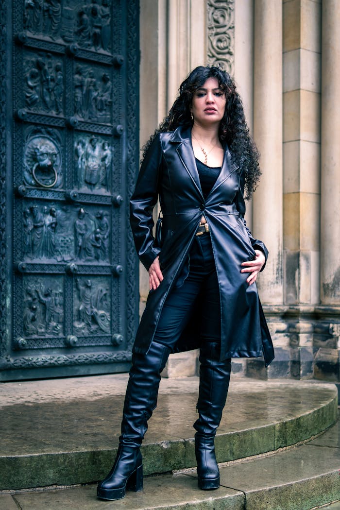 Stylish woman in black leather coat and boots poses in front of a historical door.