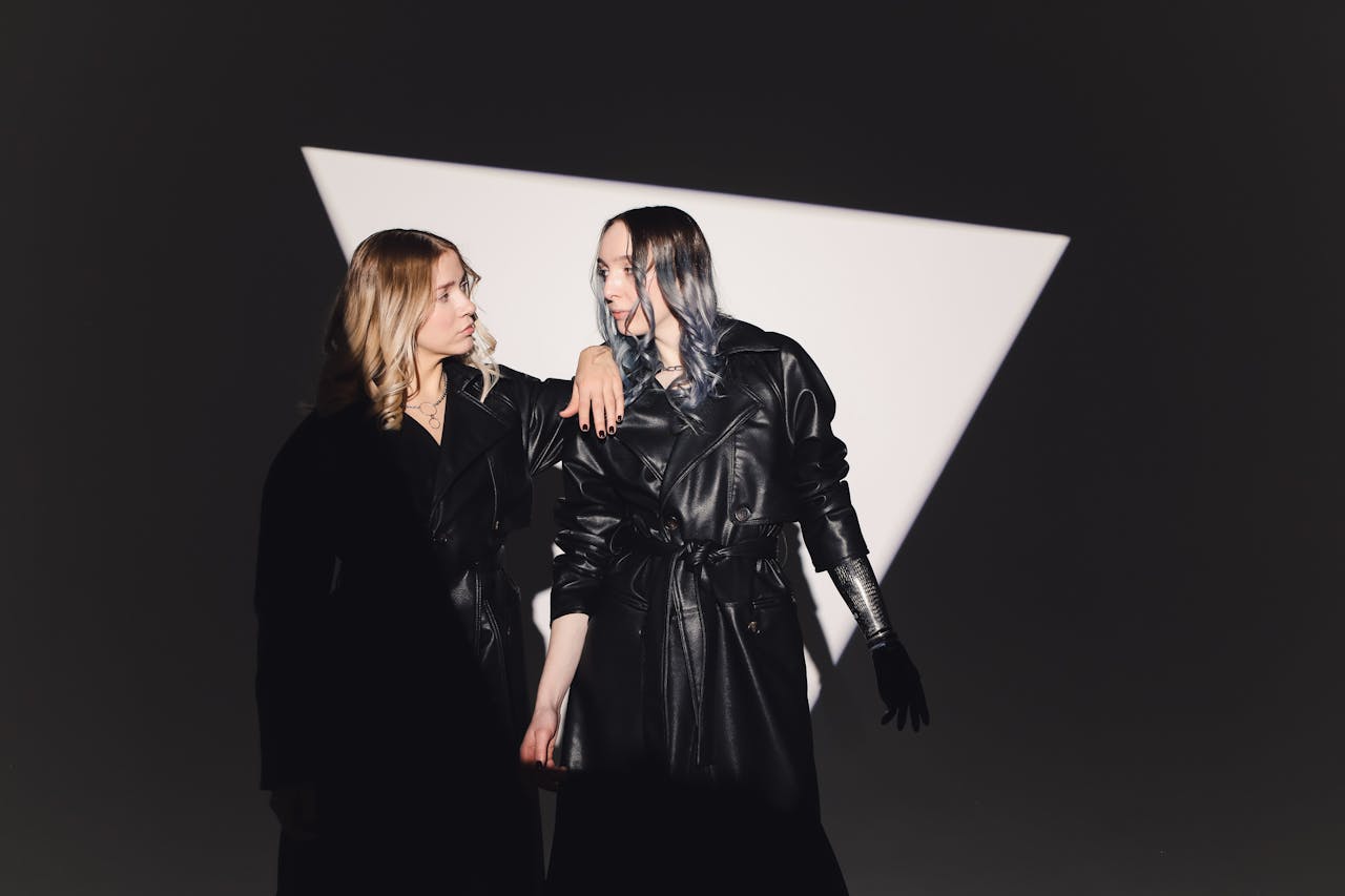 Two women in black leather coats stand confidently in a studio setting with dramatic lighting.