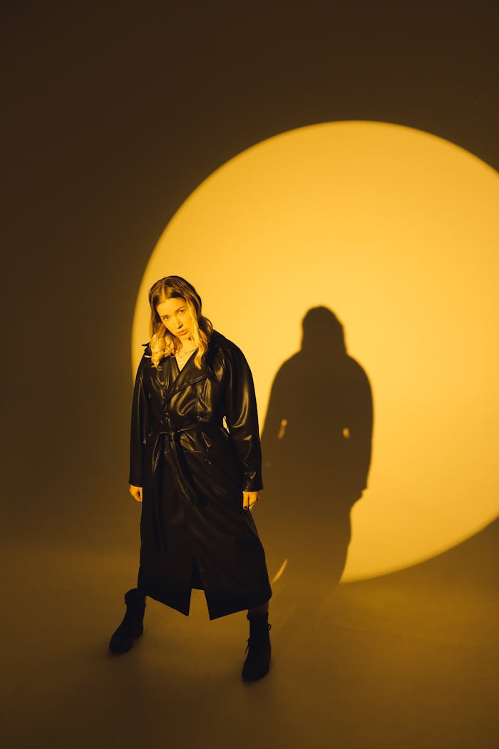 Fashionable woman wearing a black leather coat stands in dramatic lighting, casting a bold shadow.
